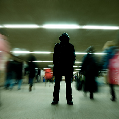 Hooded person standing in shadow while blurred people move past.